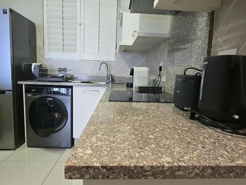 A functional kitchen featuring granite countertops, tiled backsplash, built-in cabinetry, and integrated laundry plumbing, finished with ceramic floor tiles and shutter blinds.