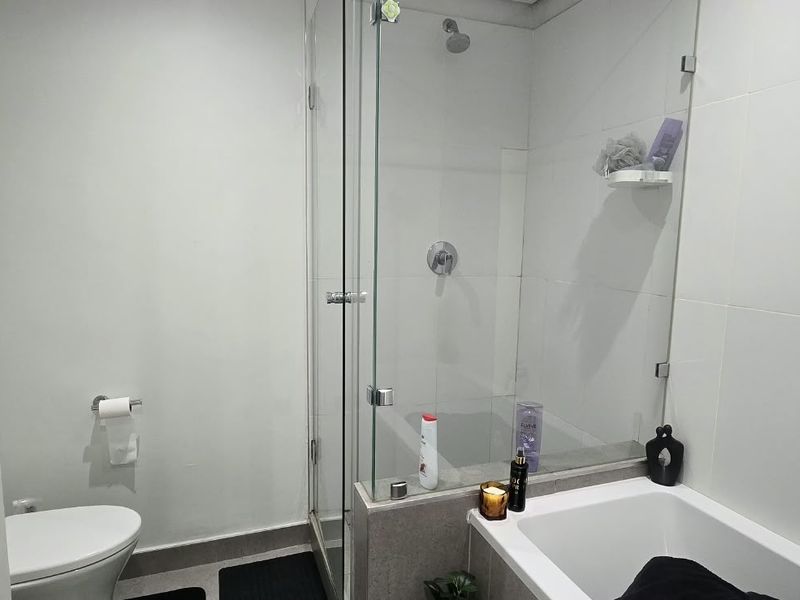 A clean, modern bathroom featuring a combined bathtub and shower enclosure with glass screen, white wall tiling, and a standard toilet.
