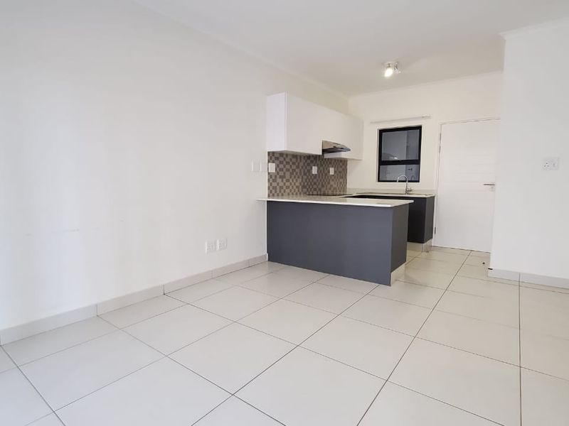 A bright, modern open-plan living and kitchen area featuring neutral tiled flooring, contemporary cabinetry, and a functional kitchen island with a mosaic backsplash.