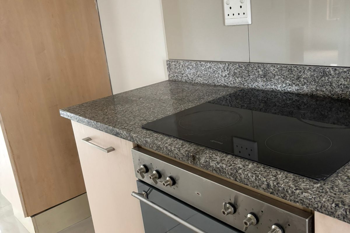 A functional kitchen area featuring granite countertops, a built-in oven with electric hob, extractor fan, and light-toned cabinetry with tiled flooring.