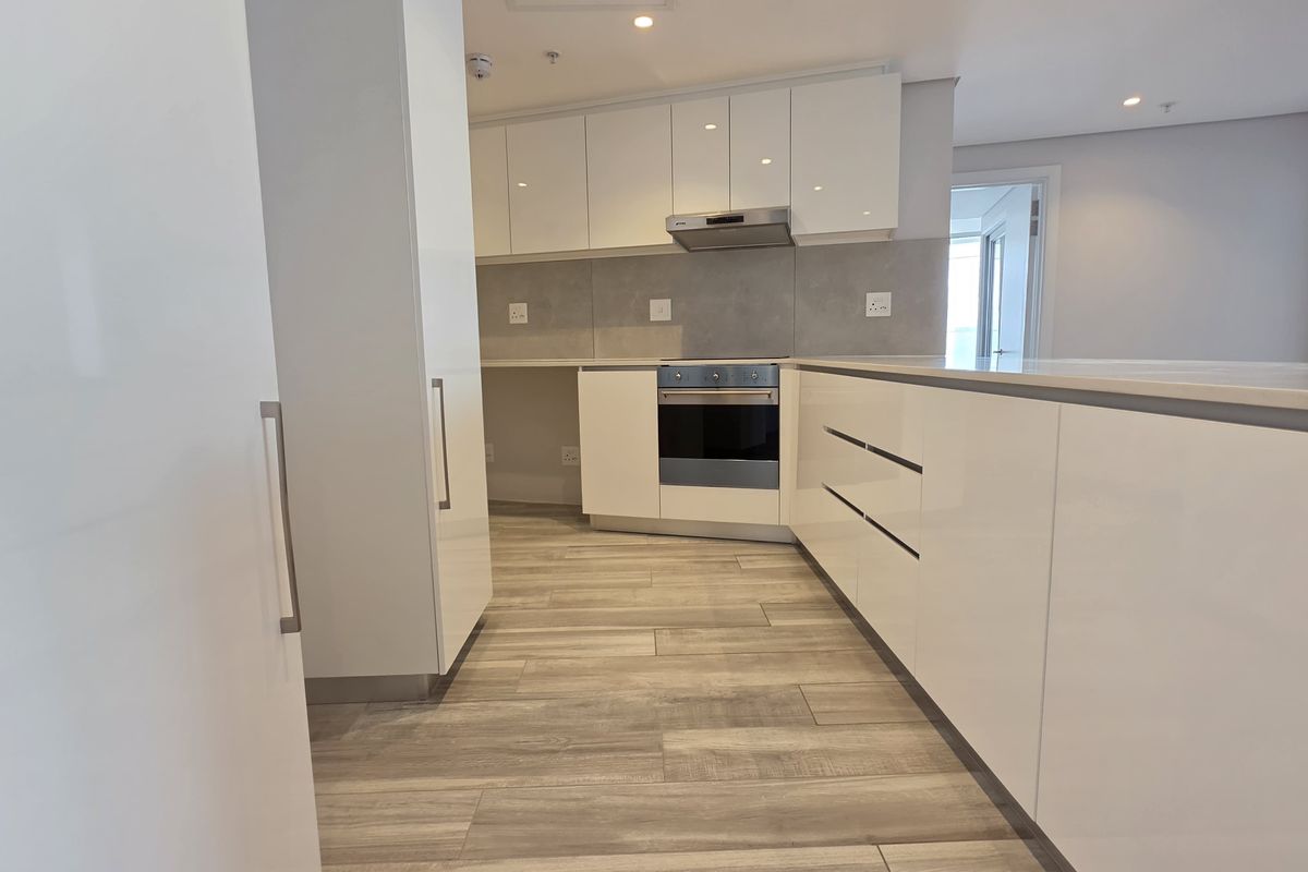 Modern kitchen featuring sleek white gloss cabinetry, stone countertops, built-in oven, extractor fan, and durable wood-look tile flooring in an open-plan layout.