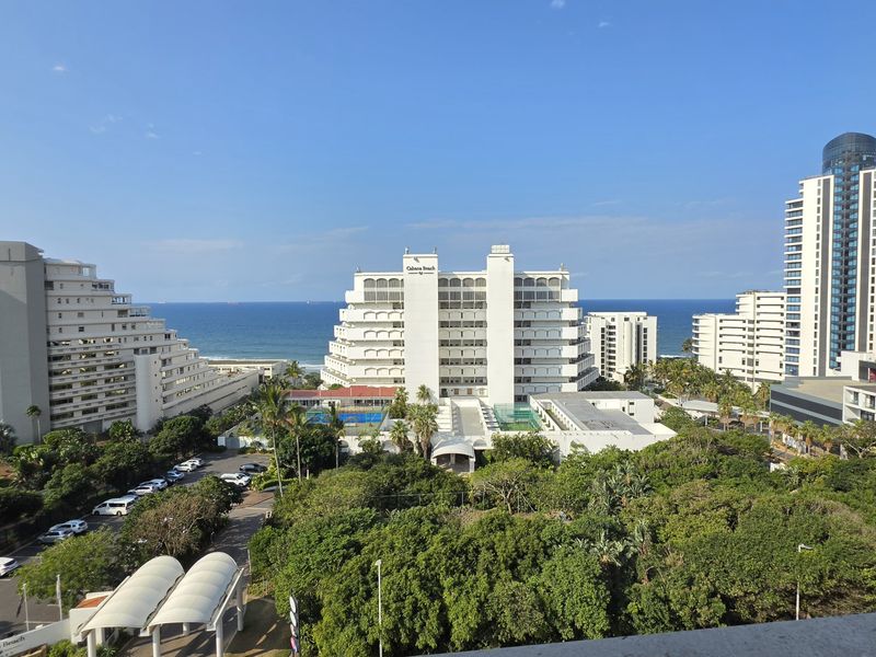 A wide-angle elevated view overlooking a coastal urban area, featuring surrounding high-rise apartment buildings, lush greenery, and a clear view of the ocean.