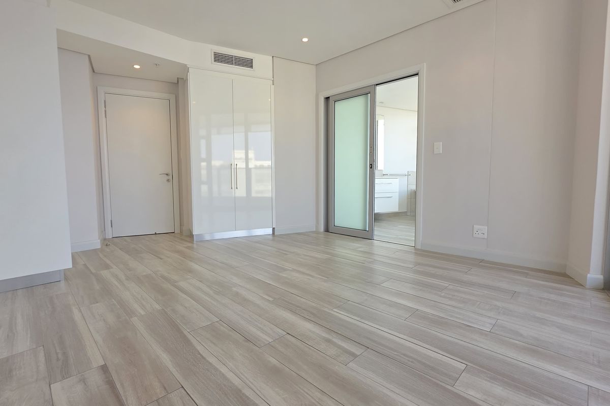 A bright, modern bedroom featuring wood-look tile flooring, built-in cupboards, and a sliding glass door leading to an en-suite bathroom.