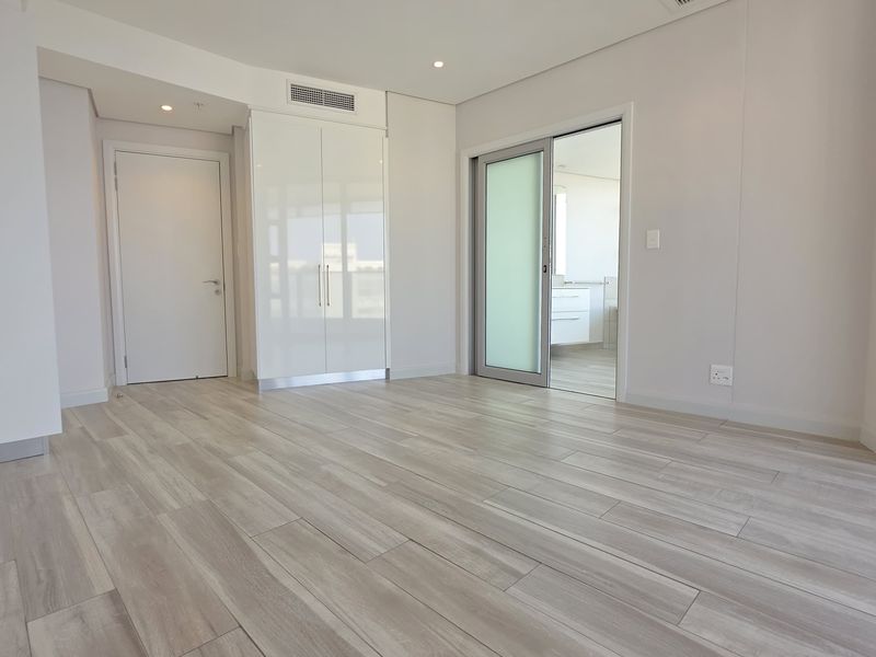 A bright, modern bedroom featuring wood-look tile flooring, built-in cupboards, and a sliding glass door leading to an en-suite bathroom.
