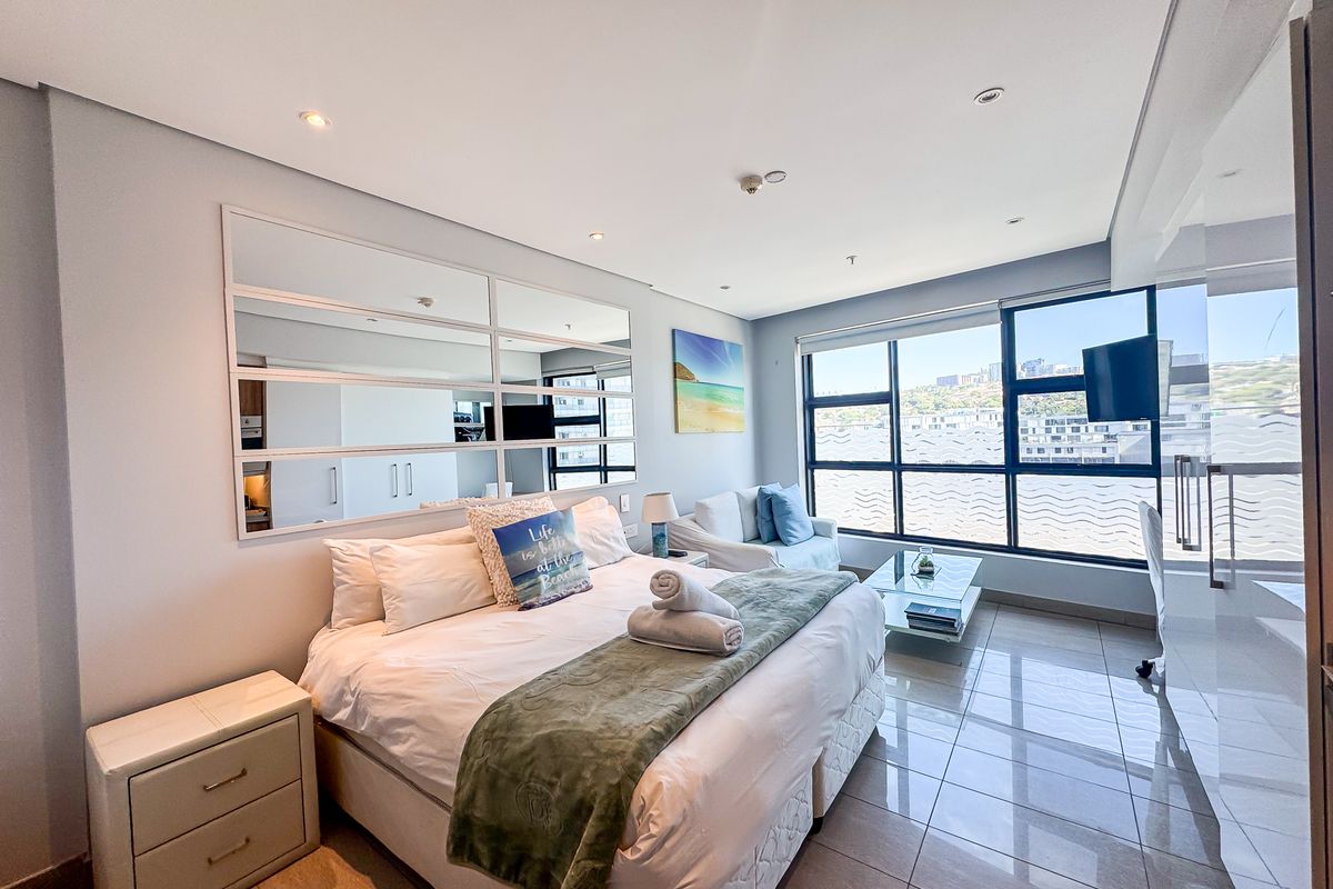 A bright and modern bedroom featuring large windows with city views, tiled flooring, recessed lighting, and a decorative mirrored wall panel.