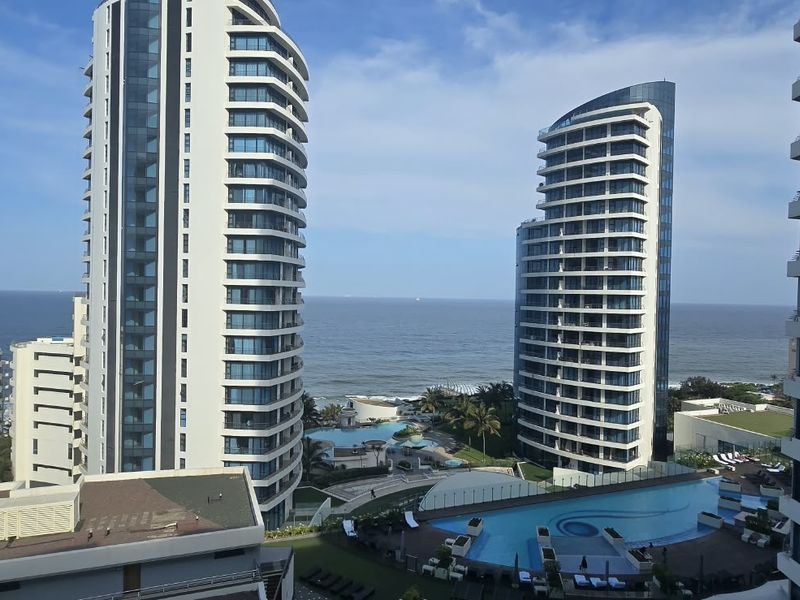 Elevated view overlooking modern high-rise apartment towers, a swimming pool area, and the ocean coastline.