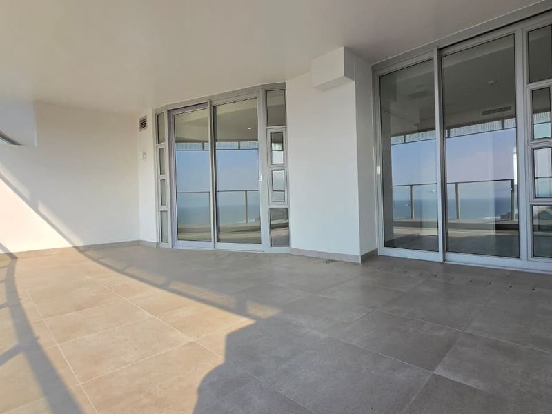 A spacious, modern covered balcony featuring large format floor tiles, glass sliding doors, and a glass balustrade with clear views.