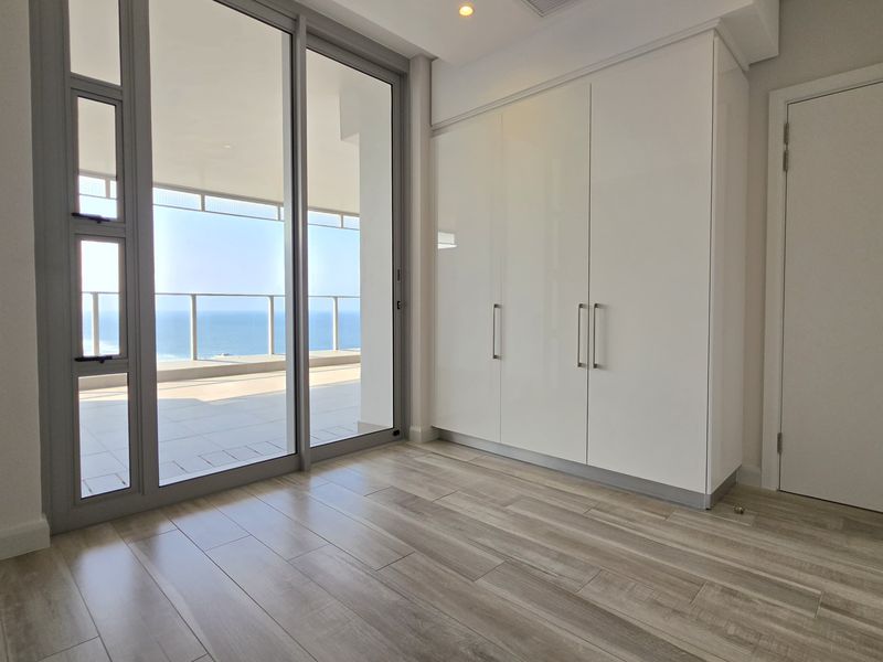 A bright, modern bedroom featuring built-in cupboards, wood-look tiled flooring, and large sliding glass doors opening onto a spacious balcony with ocean views.