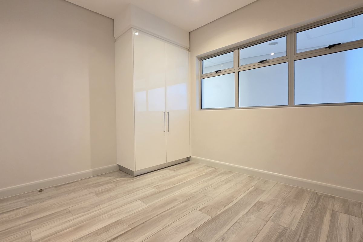 A bright, modern bedroom featuring neutral finishes, light wood-look laminate flooring, and a sleek built-in wardrobe with ample natural light.