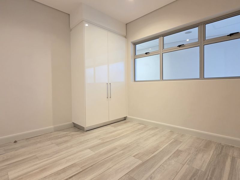 A bright, modern bedroom featuring neutral finishes, light wood-look laminate flooring, and a sleek built-in wardrobe with ample natural light.