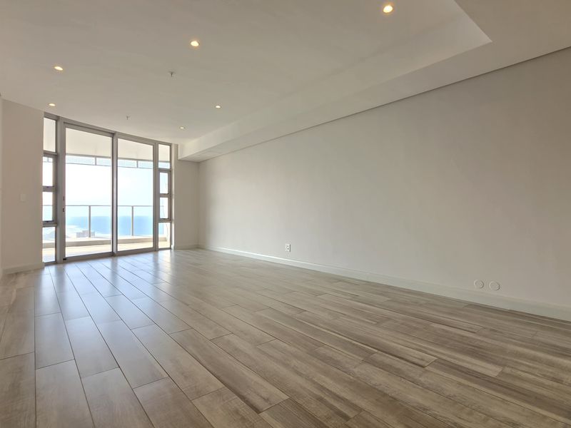 A bright, spacious, and empty living area featuring modern wood-look tile flooring, recessed ceiling lighting, and large sliding glass doors leading to a balcony.