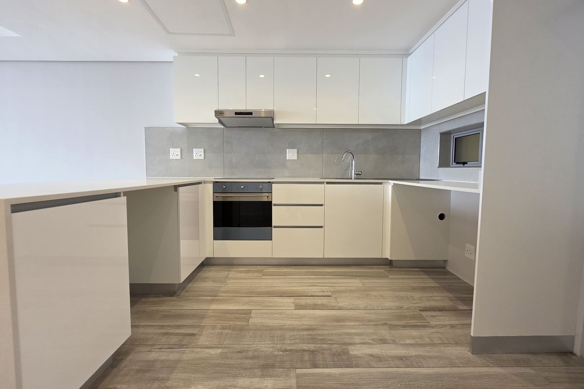 Modern, minimalist kitchen featuring high-gloss white cabinetry, stone countertops, integrated oven, extractor fan, and wood-look flooring in a clean, contemporary layout.