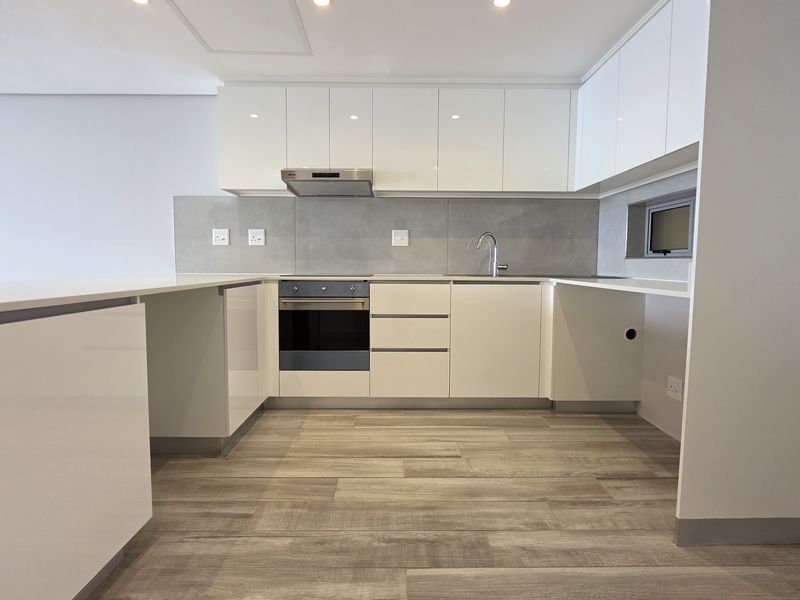 Modern, minimalist kitchen featuring high-gloss white cabinetry, stone countertops, integrated oven, extractor fan, and wood-look flooring in a clean, contemporary layout.