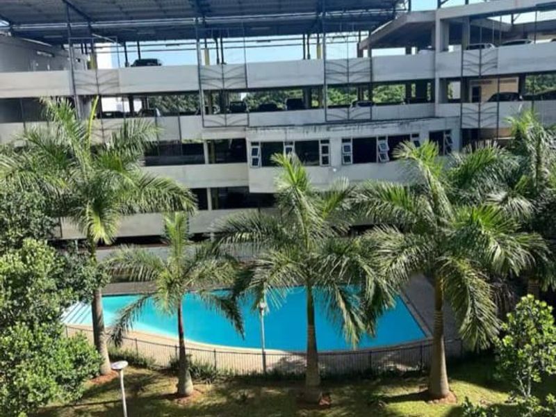 View of a communal swimming pool set within a landscaped garden area, adjacent to a multi-level parking structure.