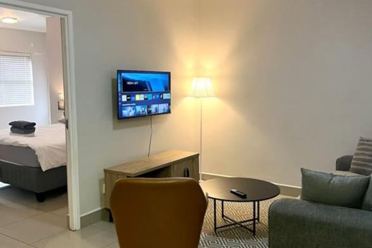 A well-maintained living room featuring neutral tiled flooring, cove ceiling detailing, and a modern flush mount light fixture, with an adjacent bedroom visible.