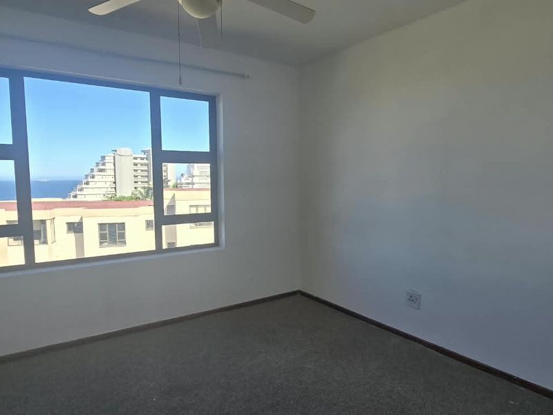 A bright, empty bedroom featuring carpeted flooring, a ceiling fan, and large windows offering an urban and coastal view.