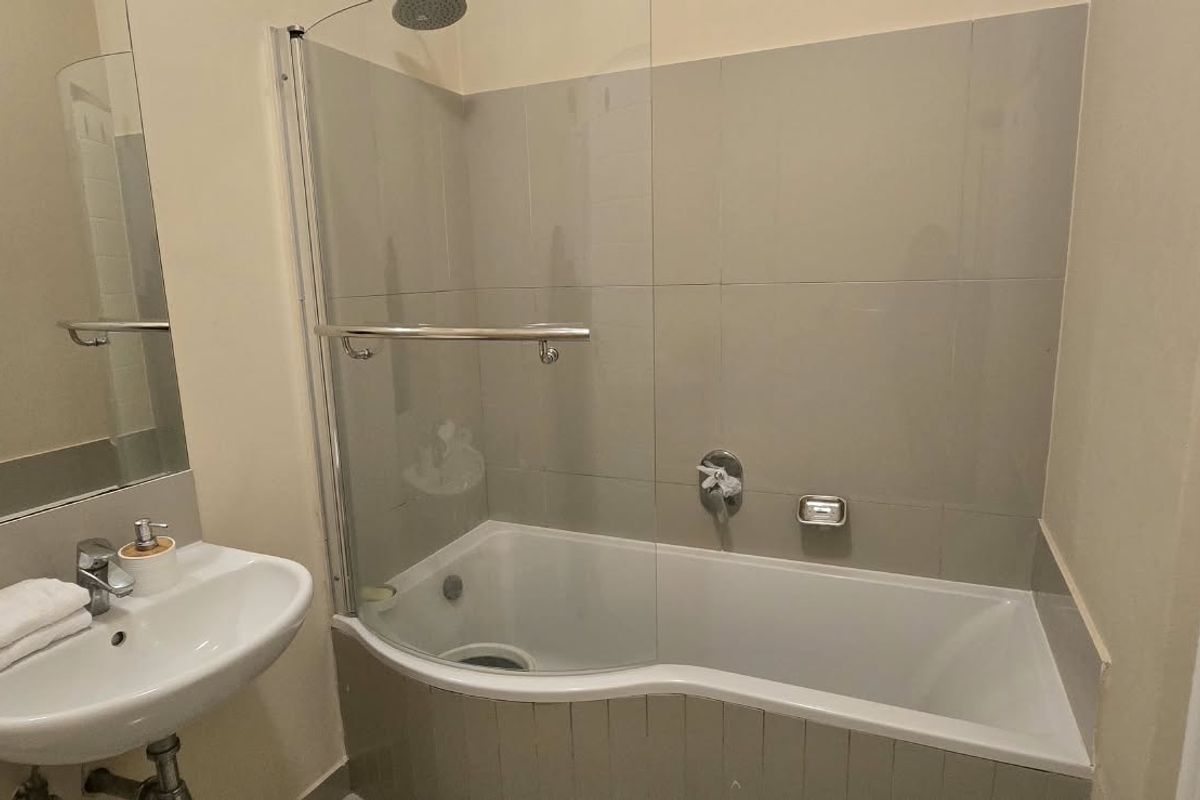 A clean, functional bathroom featuring a wall-mounted basin, a shower-over-bath combination with a glass screen, and neutral wall tiling.