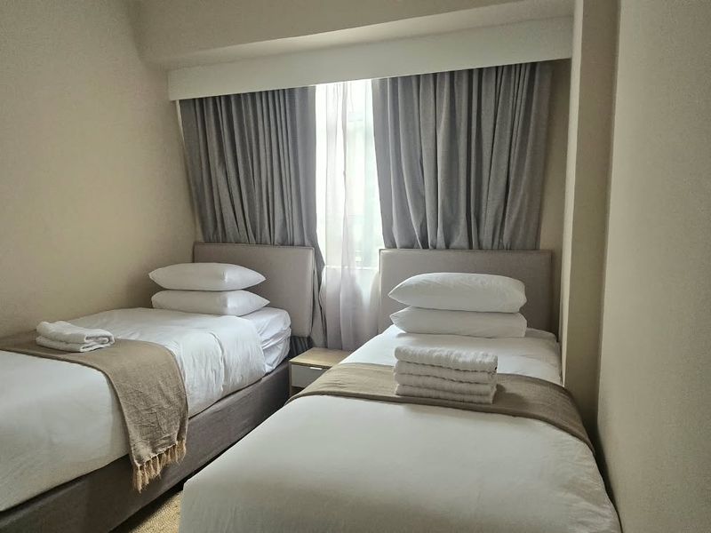 A compact bedroom featuring a neutral color palette, recessed window area with fitted curtains, and space for two single beds.