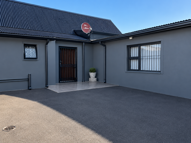 Modern exterior front facade featuring a dark grey finish, corrugated metal roof, secure entrance with security gate, and a clean asphalt driveway.