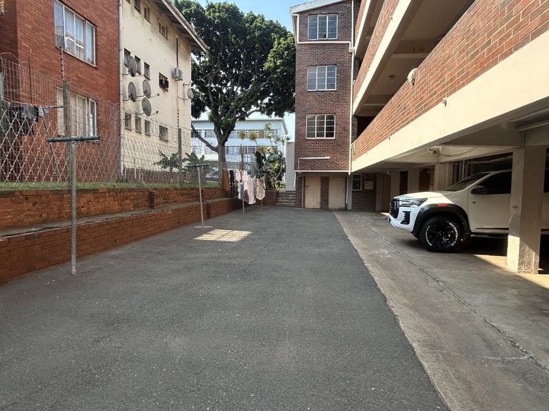 Exterior view of an apartment complex featuring a paved driveway, covered parking area, and brick building facades with security fencing.