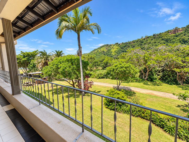 A spacious covered balcony featuring a metal balustrade and tiled flooring, offering scenic views of the surrounding garden and lush hillside landscape.