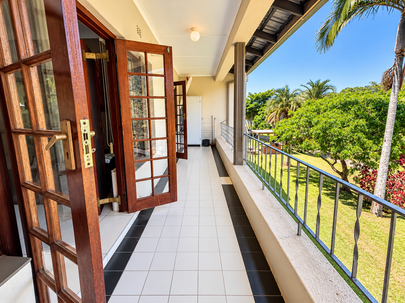 A spacious covered balcony featuring tiled flooring, a metal balustrade, and timber-framed glass doors, offering a pleasant view of the surrounding garden.