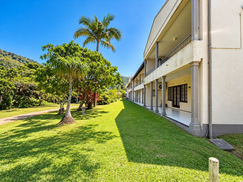 Exterior view of an apartment building featuring a covered ground-floor patio, upper-level balcony with railings, and a well-maintained garden area.