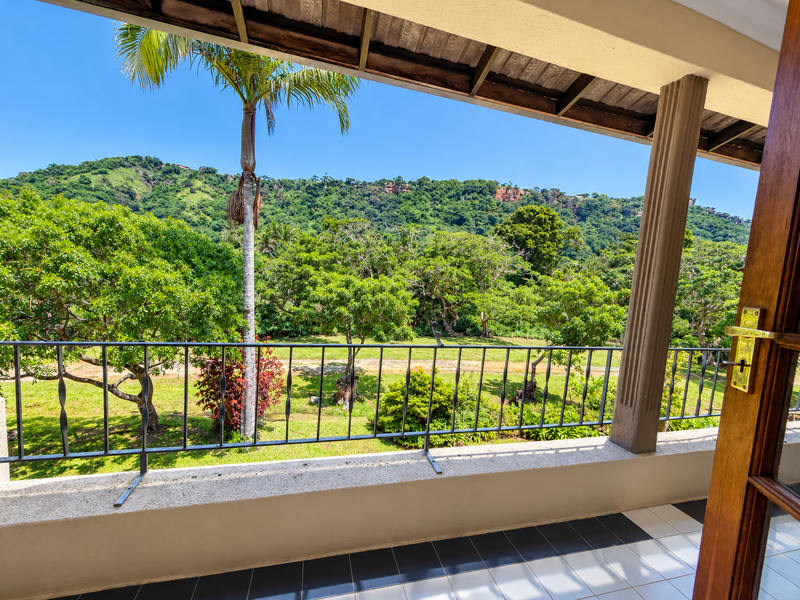 A covered balcony featuring tiled flooring, a decorative fluted column, and wrought iron railings, offering expansive views of the surrounding lush green landscape.