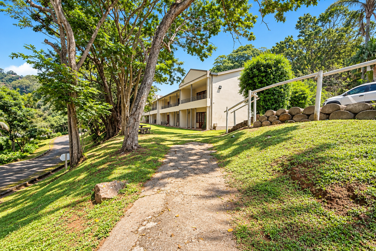 A view of the apartment building exterior featuring a concrete pathway, landscaped grounds, and rear-facing balconies set against a lush, tree-lined backdrop.