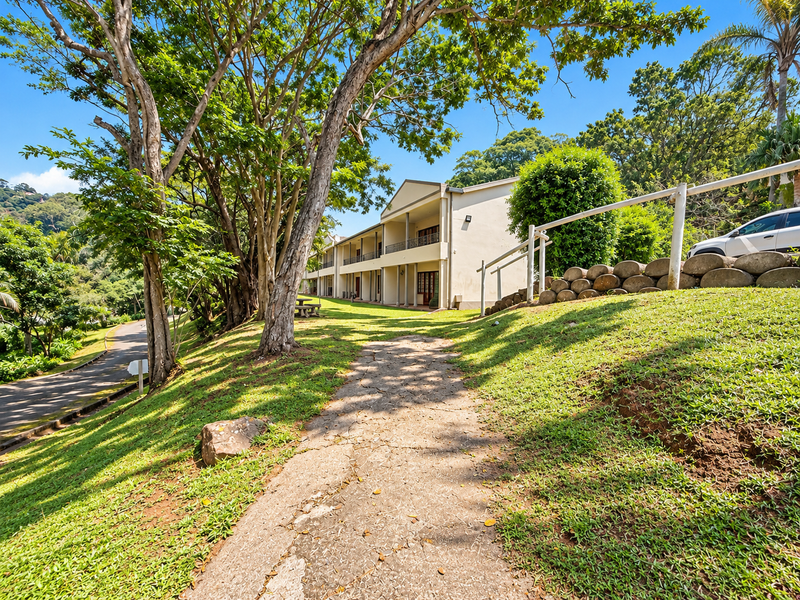 A view of the apartment building exterior featuring a concrete pathway, landscaped grounds, and rear-facing balconies set against a lush, tree-lined backdrop.
