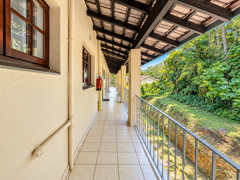 A covered outdoor balcony featuring tiled flooring, a metal balustrade, and a timber roof structure, overlooking a lush green garden area.