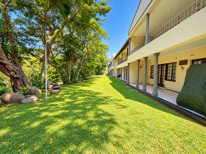 A well-maintained rear exterior view of the apartment complex featuring a spacious covered patio, tiled flooring, and a lush, tree-lined garden area.