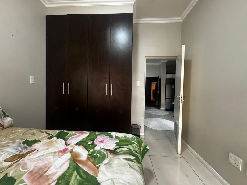 A bedroom featuring dark wood built-in cupboards, tiled flooring, and decorative ceiling cornicing, with an open doorway leading to an adjacent living area.
