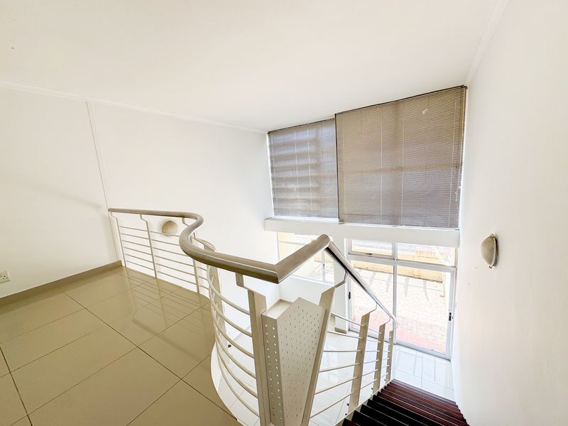 A bright, open-plan landing area featuring a curved metal staircase railing, tiled flooring, and large windows that provide ample natural light.