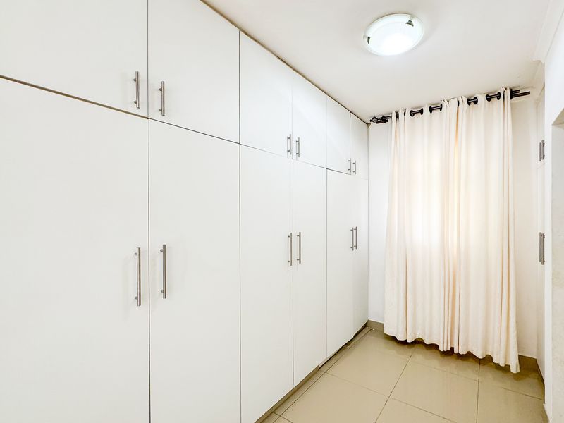 A clean, empty bedroom featuring extensive white built-in cupboards, neutral tiled flooring, and a simple ceiling light fixture.