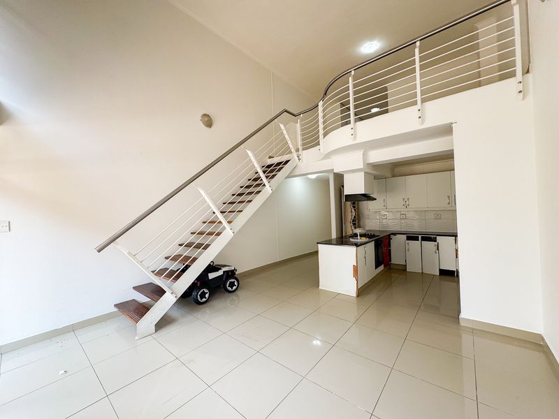 Open-plan living area featuring a modern staircase leading to a mezzanine level, adjacent to a functional kitchen with built-in cabinetry and tiled flooring.
