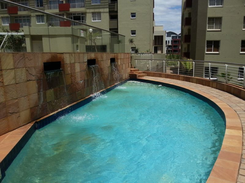 A communal swimming pool area featuring a stone-clad water feature, glass balustrades, and paved decking within an apartment complex.