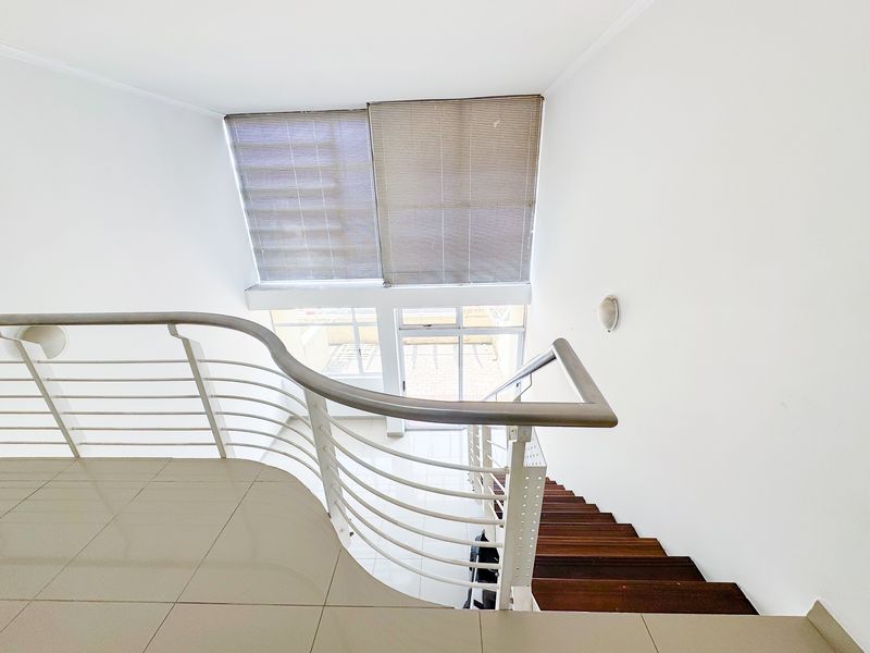 A bright, modern staircase area featuring wooden treads, a sleek metal balustrade, and large windows with venetian blinds, leading to a tiled lower level.
