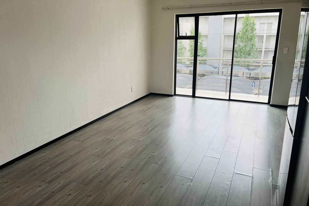 A bright, empty living area featuring modern laminate flooring and a large sliding glass door providing access to a balcony.