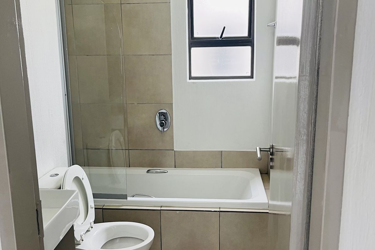 A clean, functional bathroom featuring a built-in bathtub with a glass shower screen, a wall-mounted basin, and neutral floor-to-ceiling tiling.