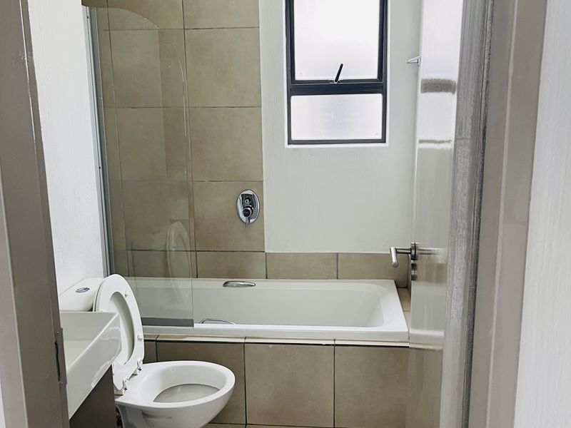 A clean, functional bathroom featuring a built-in bathtub with a glass shower screen, a wall-mounted basin, and neutral floor-to-ceiling tiling.