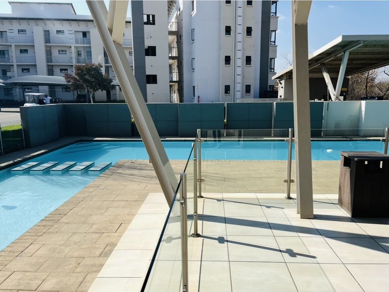 A well-maintained communal swimming pool area featuring a paved deck, glass balustrades, and modern steel structural supports within an apartment complex.