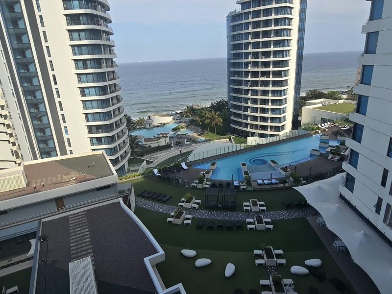 Elevated view overlooking the apartment complex, featuring a swimming pool, landscaped garden areas, and direct ocean views.