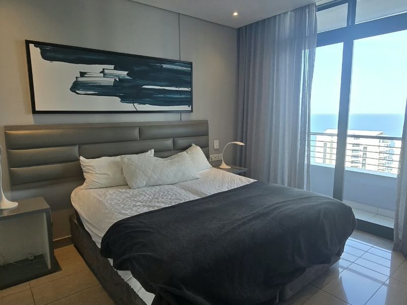 A modern bedroom featuring tiled flooring, a built-in headboard, and large sliding glass doors that open to a balcony with ocean views.