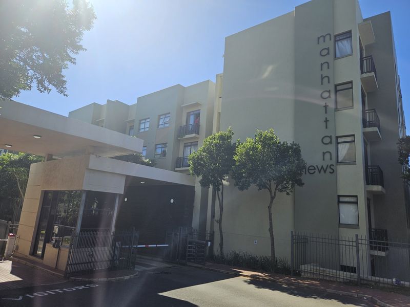Front exterior view of Manhattan Mews apartment complex featuring a secure gated entrance, guard house, and multi-story residential building facade.