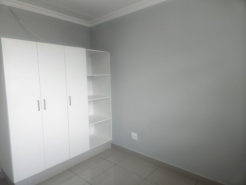 A clean, empty bedroom featuring neutral grey walls, large format floor tiles, and a modern white built-in wardrobe with integrated shelving.
