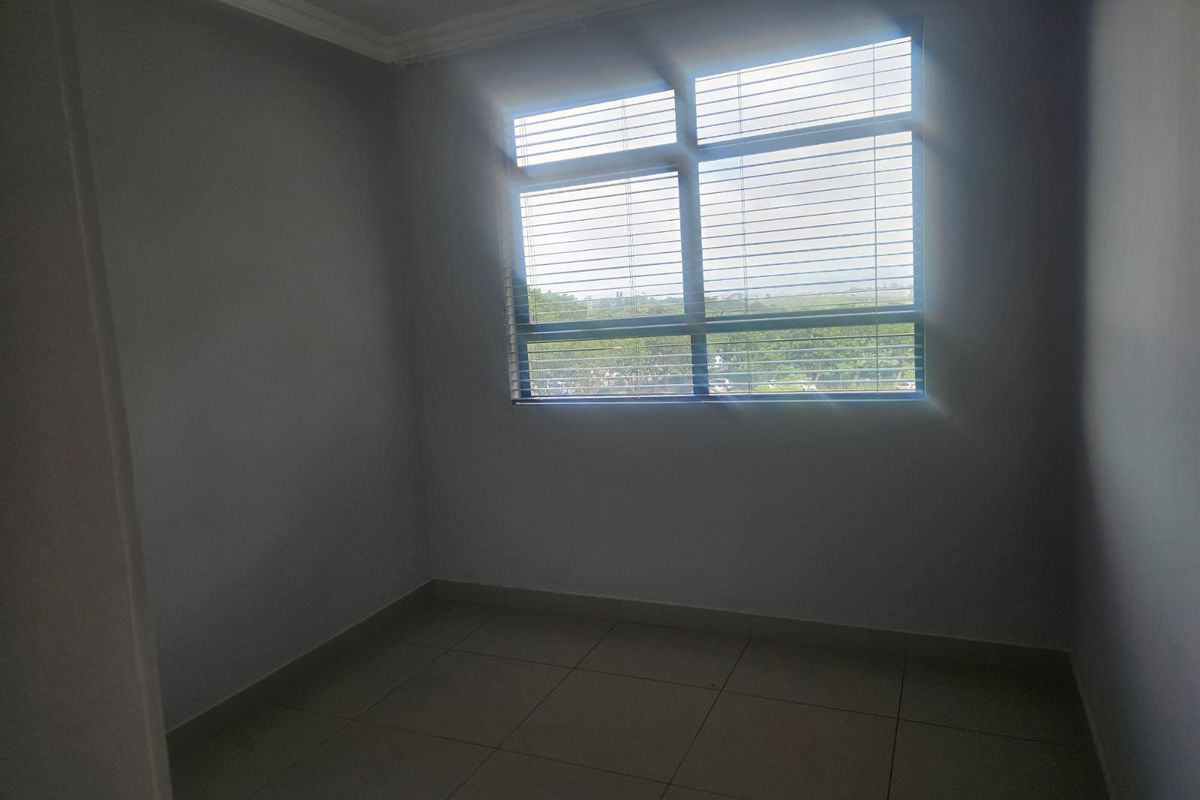 A bright, empty bedroom featuring neutral tiled flooring and a large window fitted with venetian blinds, offering a clean and versatile living space.