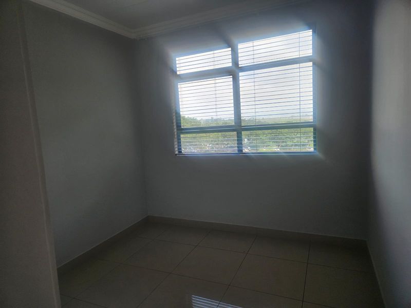A bright, empty bedroom featuring neutral tiled flooring and a large window fitted with venetian blinds, offering a clean and versatile living space.