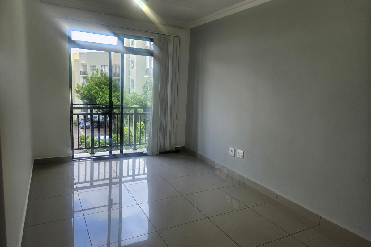 An empty, well-lit living room featuring neutral tiled flooring, cove molding, and a sliding glass door providing access to a balcony.