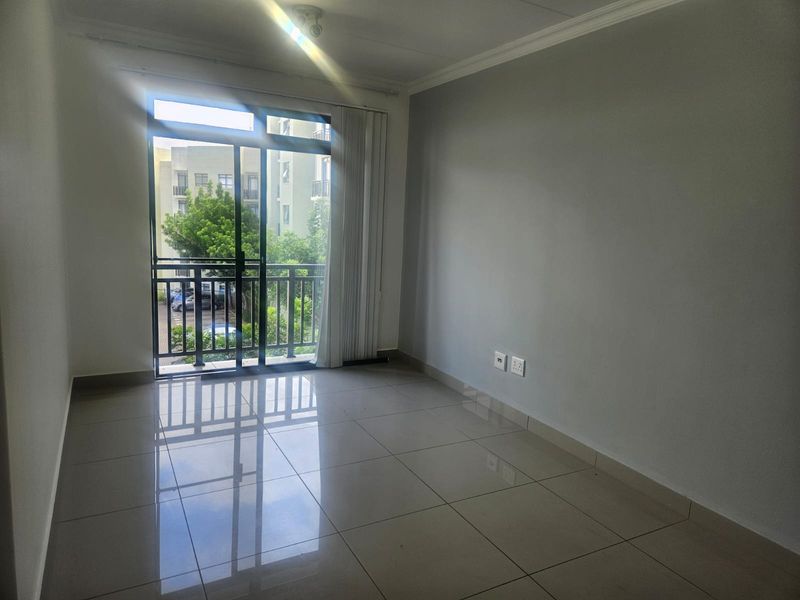 An empty, well-lit living room featuring neutral tiled flooring, cove molding, and a sliding glass door providing access to a balcony.